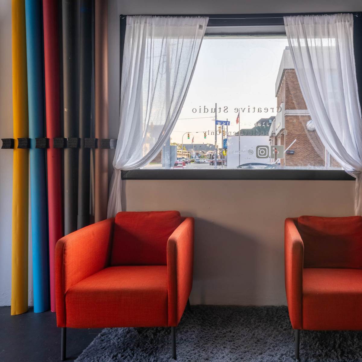 Two orange chairs are positioned in front of a large window, adorned with sheer white curtains, overlooking a street scene with colorful rolls of backdrop material displayed nearby.