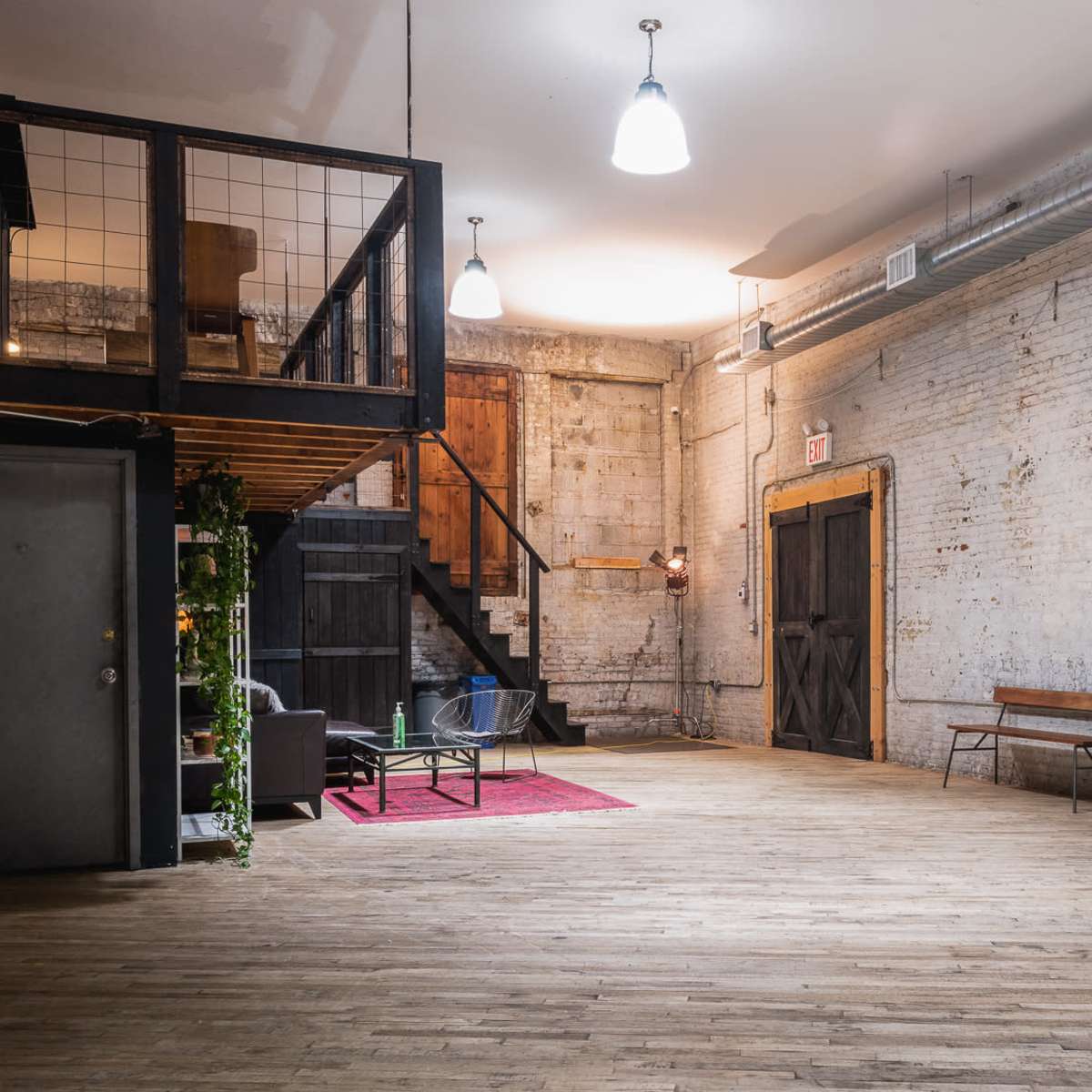 A spacious, industrial-style interior with exposed brick walls, wooden floors, a staircase, and minimal furniture.