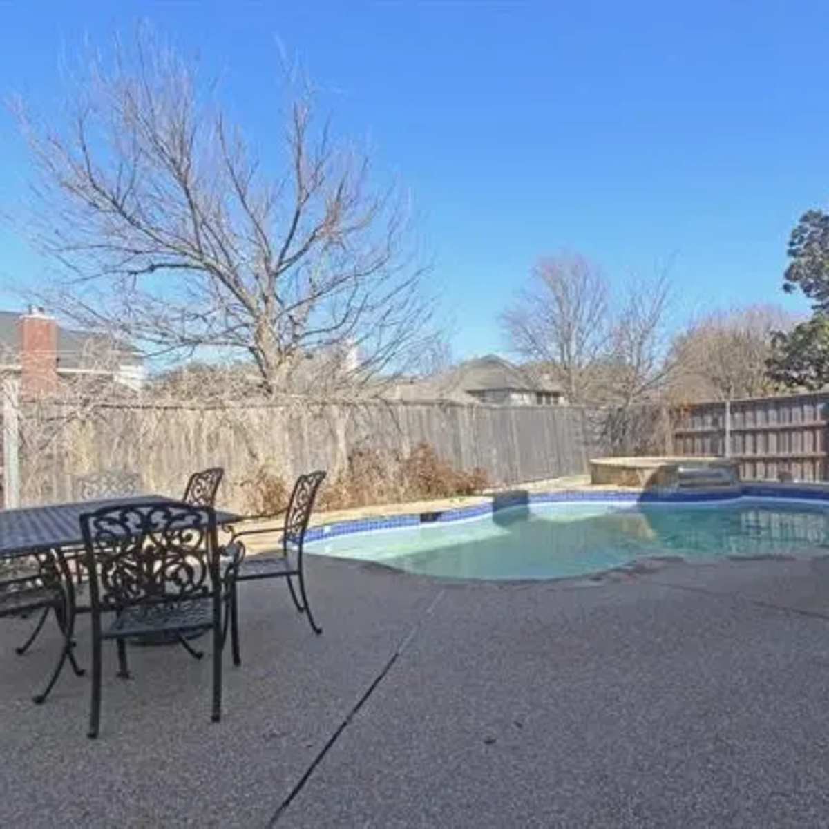A backyard with a swimming pool surrounded by a concrete patio and outdoor dining furniture.