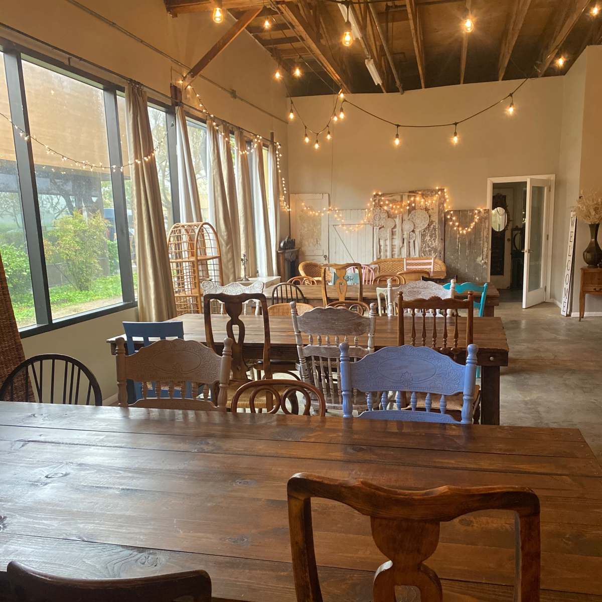 A spacious interior room with wooden tables and chairs, large windows, and string lights overhead.