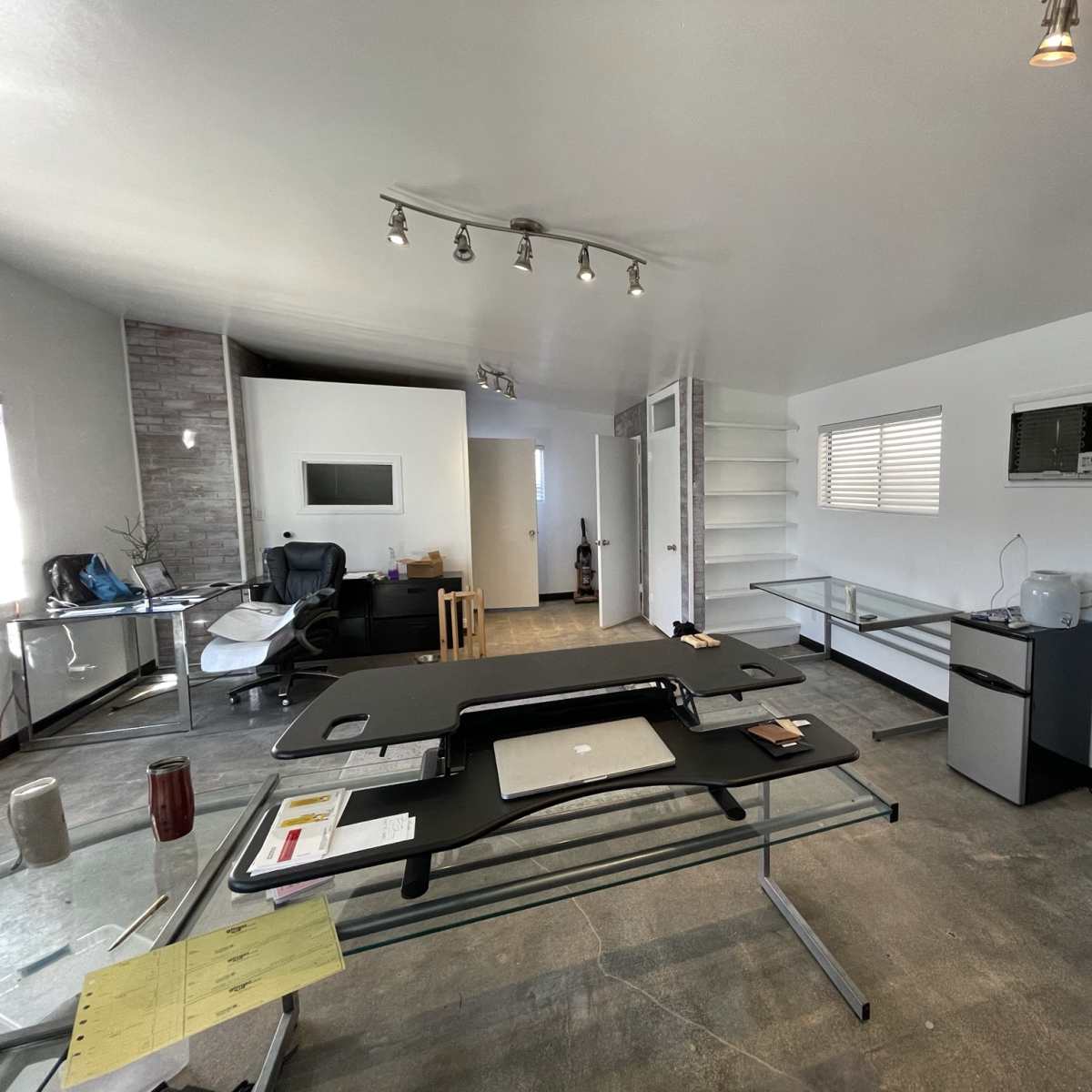 A modern, minimalistic office space with a glass desk, a chair, shelves, and an air conditioning unit.