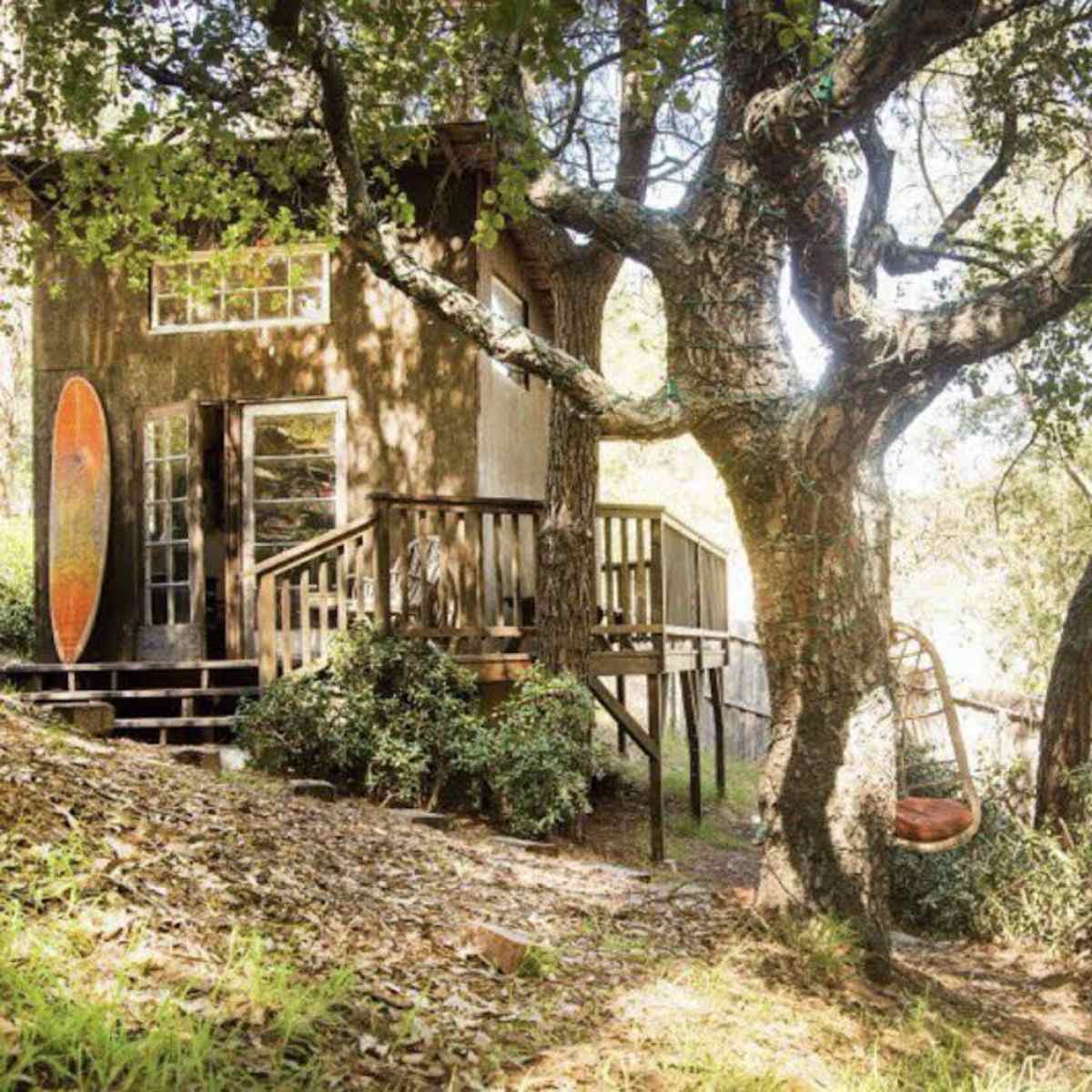 A small wooden cabin sits on a hillside, surrounded by trees, with two hanging wicker chairs and a surfboard leaning against the building.