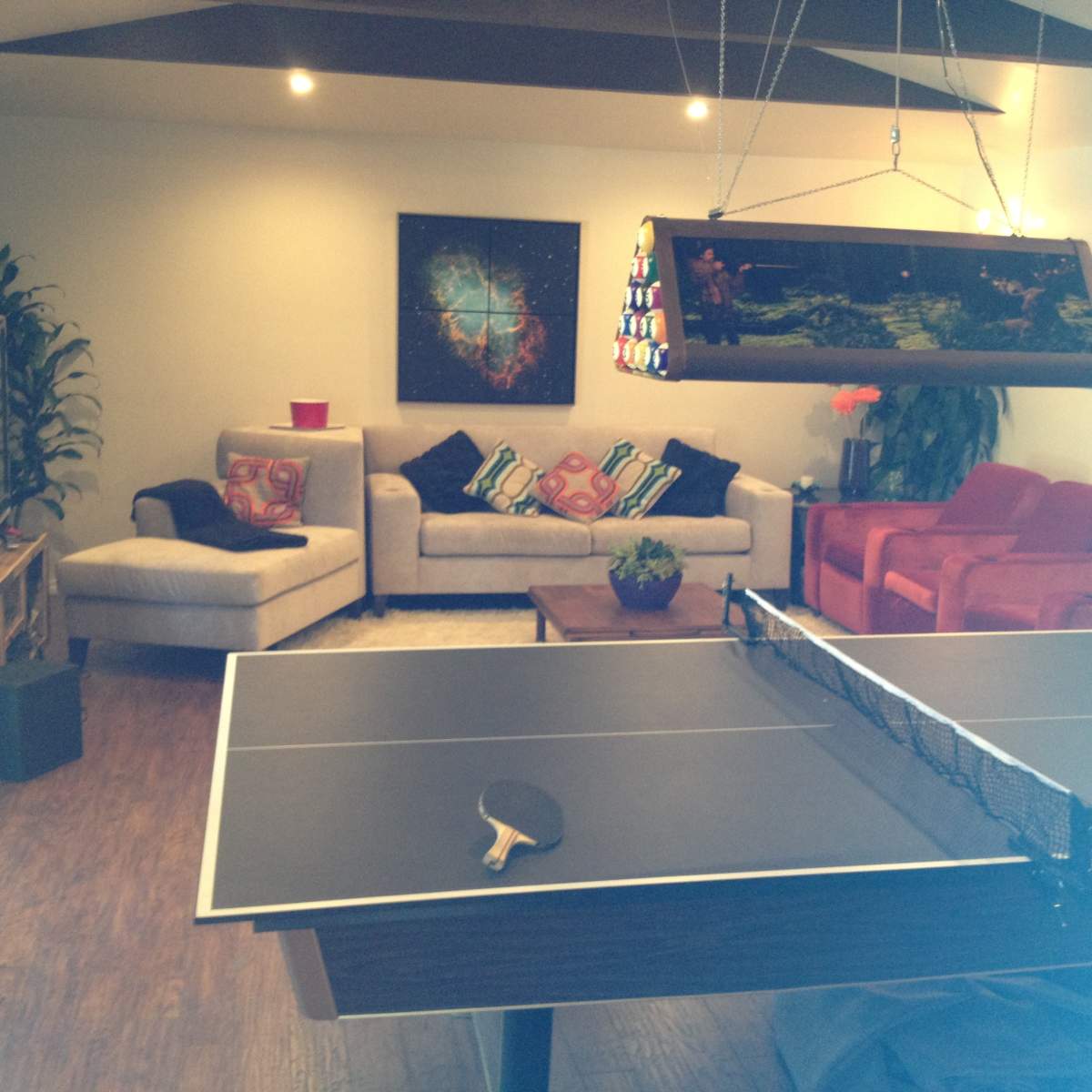 A game room features a ping pong table in the foreground, with a sofa and chairs in the background, along with decorative plants and a large light fixture.