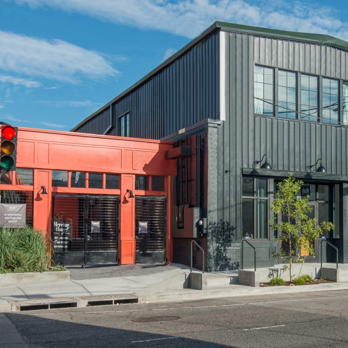 A modern building with a combination of red and dark green metal siding, featuring large windows and a small tree near the entrance.