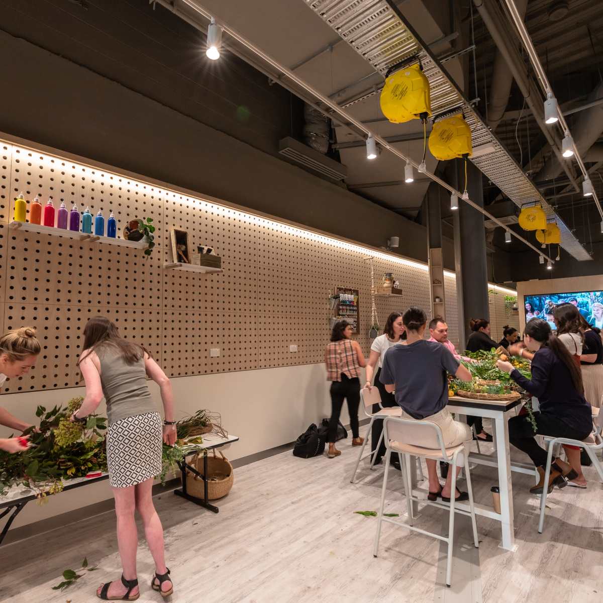 A group of people are arranging plants and foliage on tables in a well-lit, modern workspace with pegboard walls and colorful decorations.