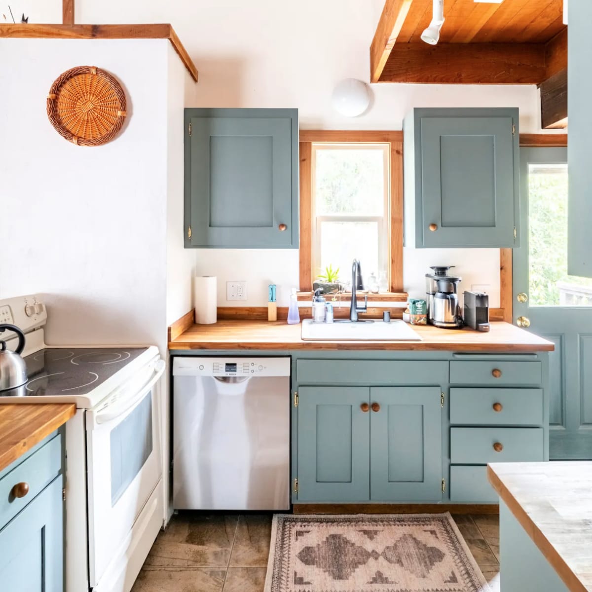 A kitchen with pale blue cabinets, a white stove, a dishwasher, and a wooden countertop, featuring natural light from a nearby window.
