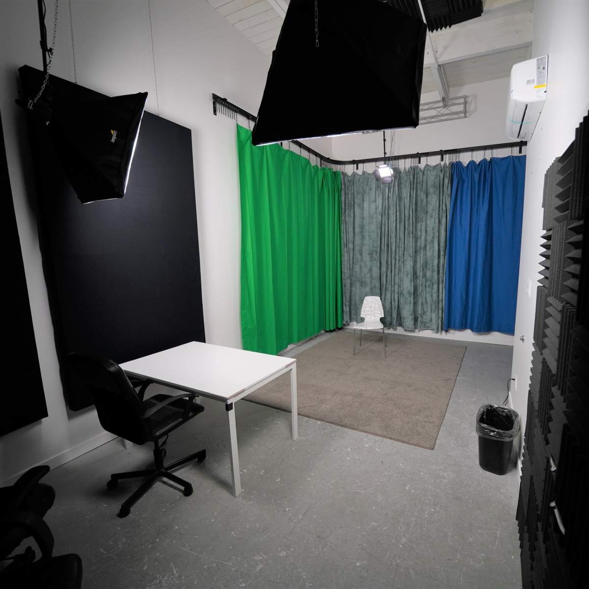 A video production studio featuring a white table and chair on a carpet, surrounded by green, blue, and gray curtains, with softbox lighting elements above.