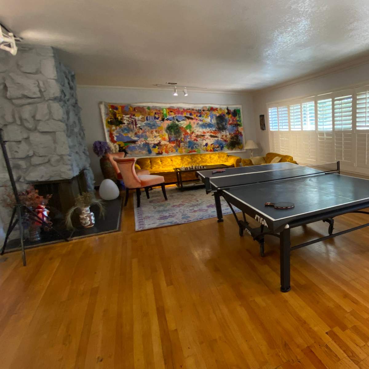 A living room featuring a ping pong table, a colorful wall mural, a yellow couch, and large windows with shutters.