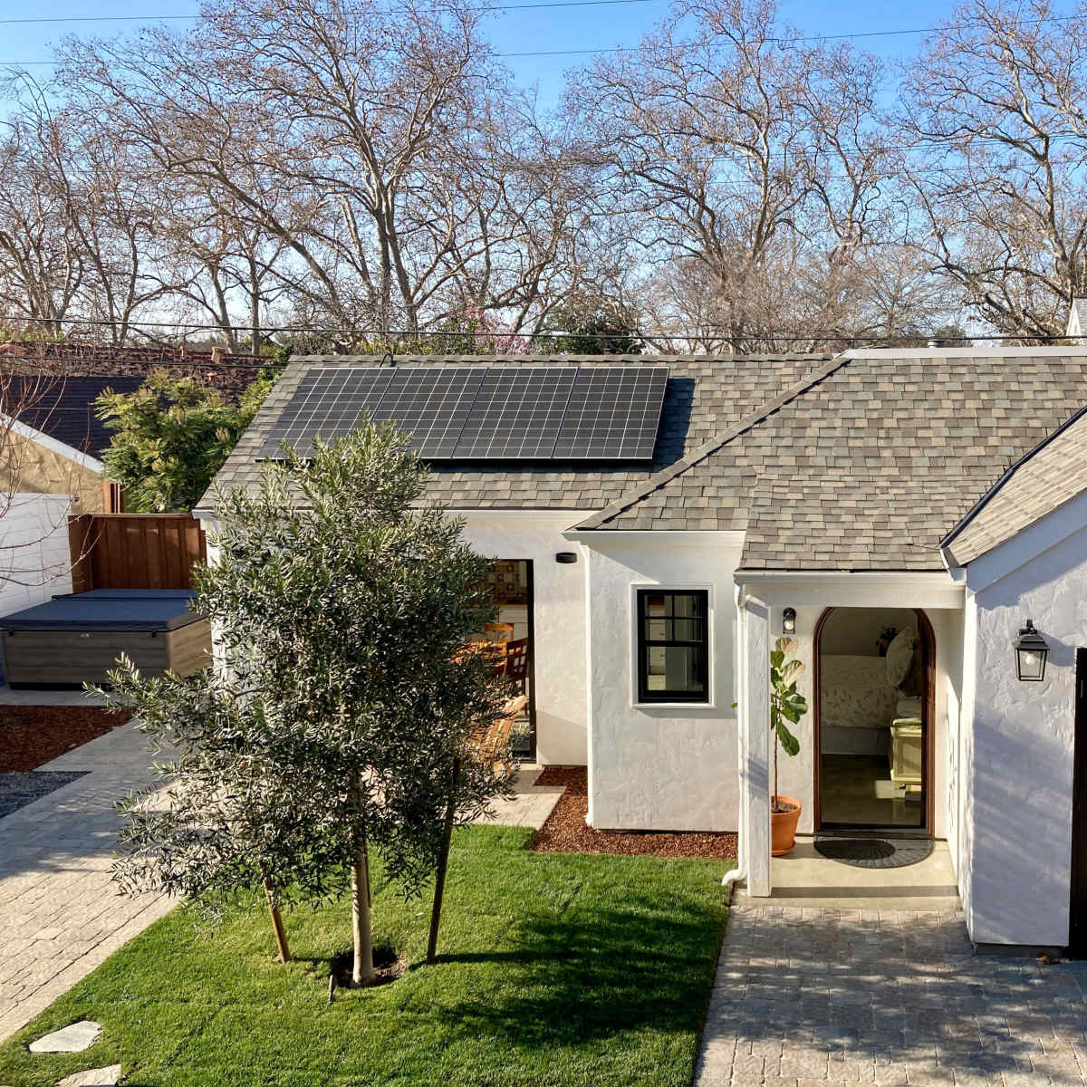 A light-colored house with a gabled roof, solar panels, and a small landscaped yard featuring an olive tree and stone pathways.