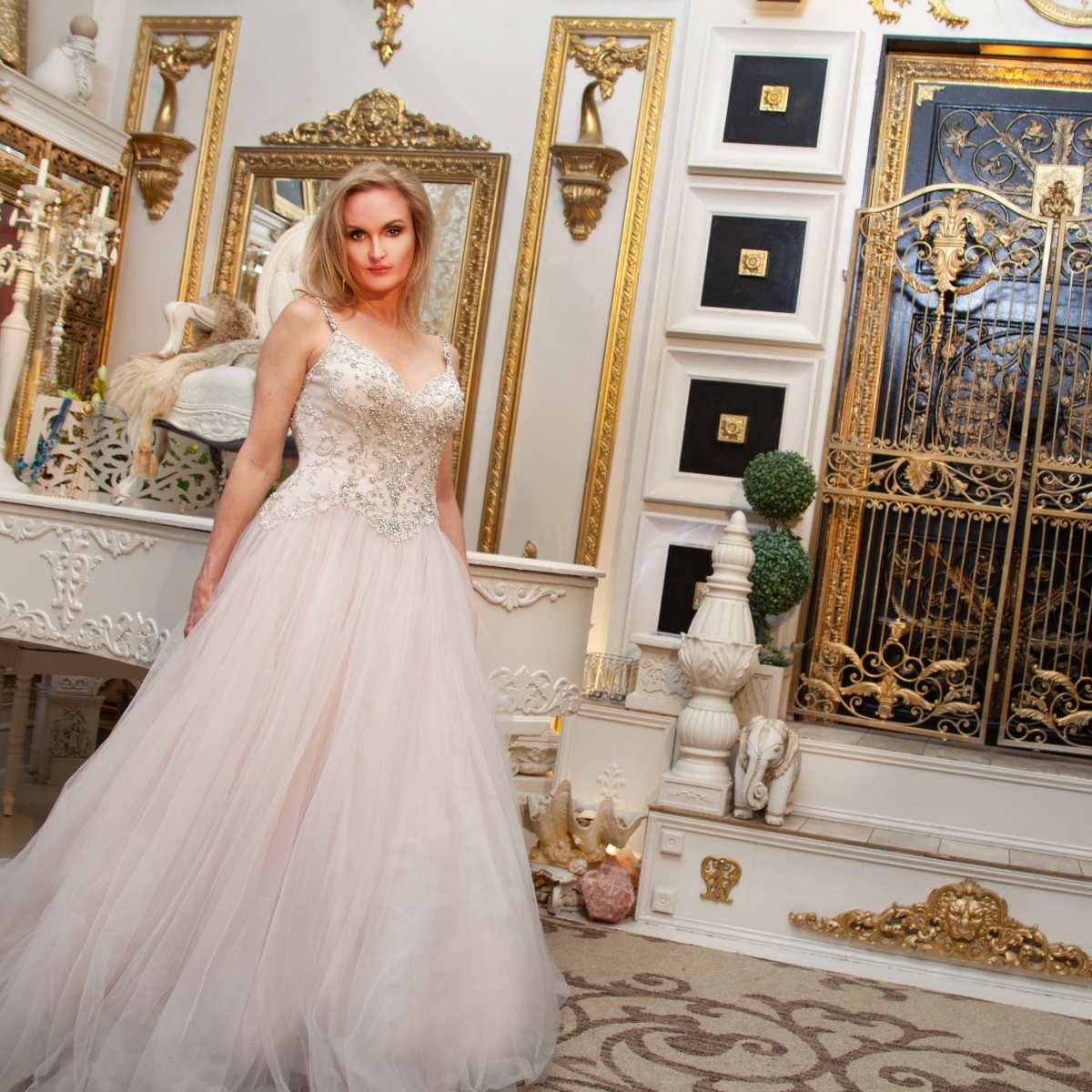 A woman in a flowing light-colored gown stands in an ornate room with intricate decorations and a gilded door.