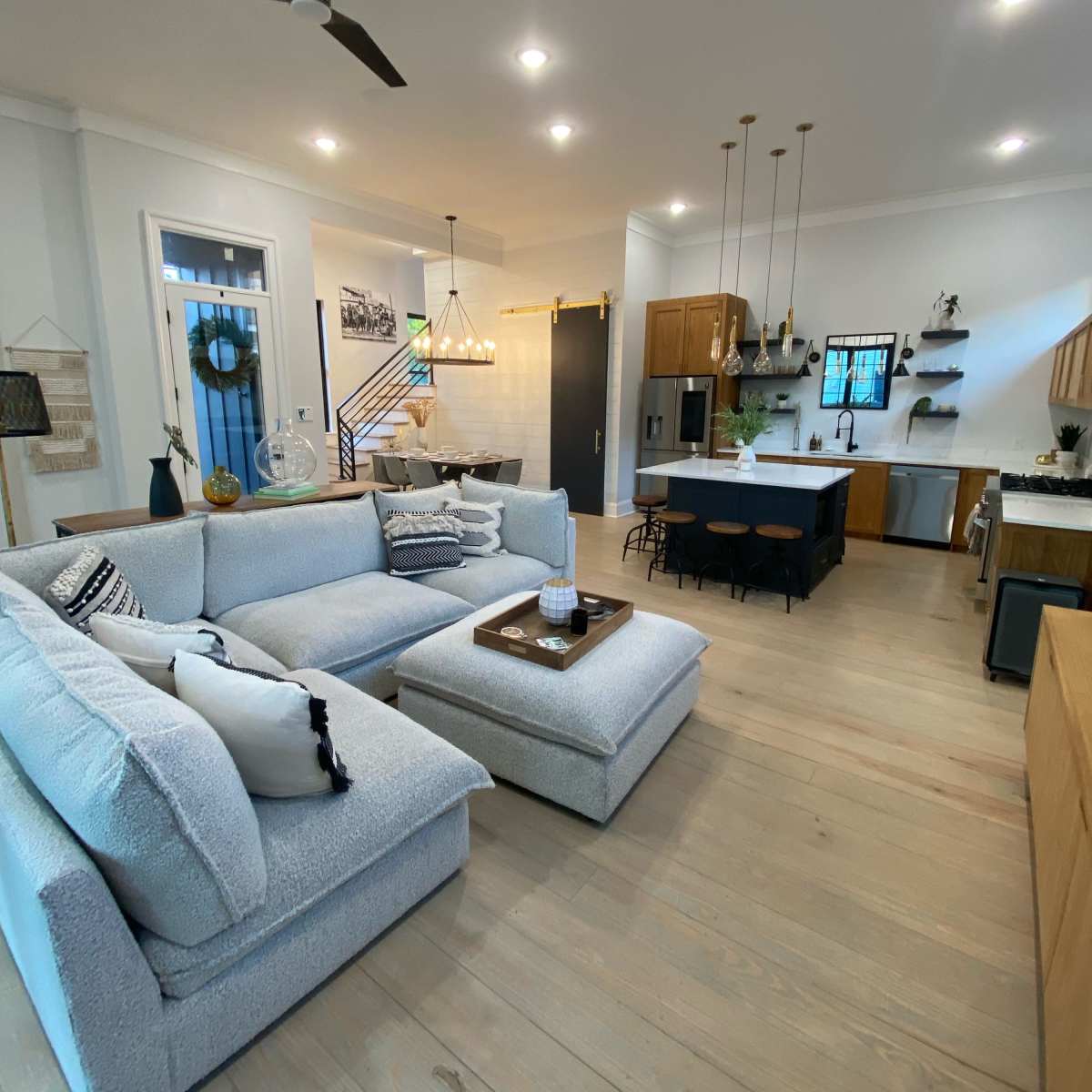 A modern living room with a light-colored sectional sofa, a coffee table, and an open kitchen area featuring wooden cabinets and a dining table.