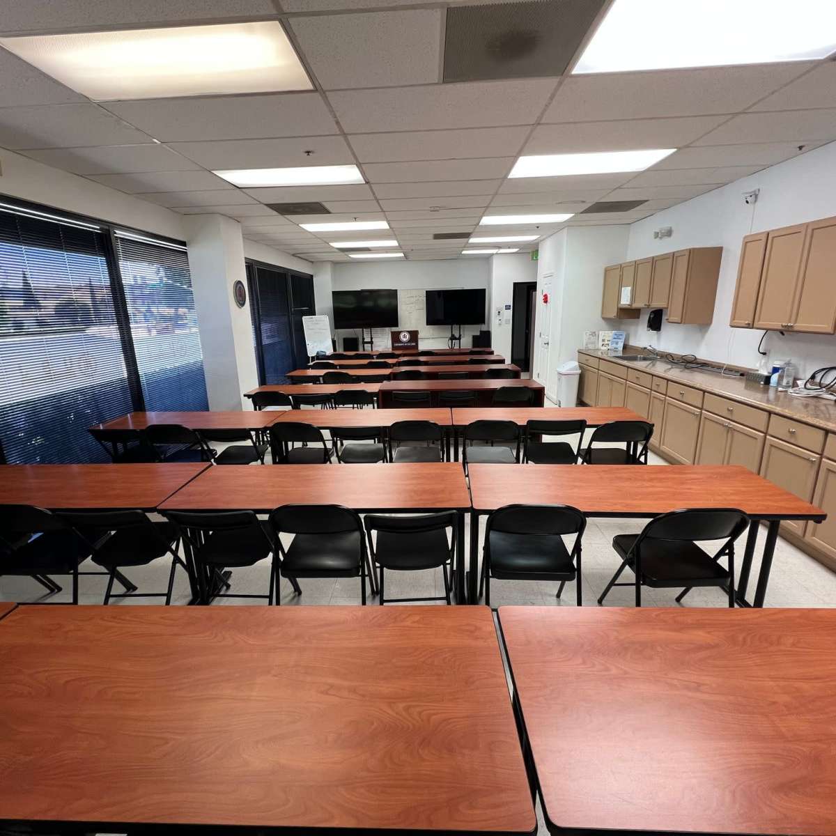 A classroom setup with rows of wooden tables and black chairs, along with a kitchenette and a large window at one end.