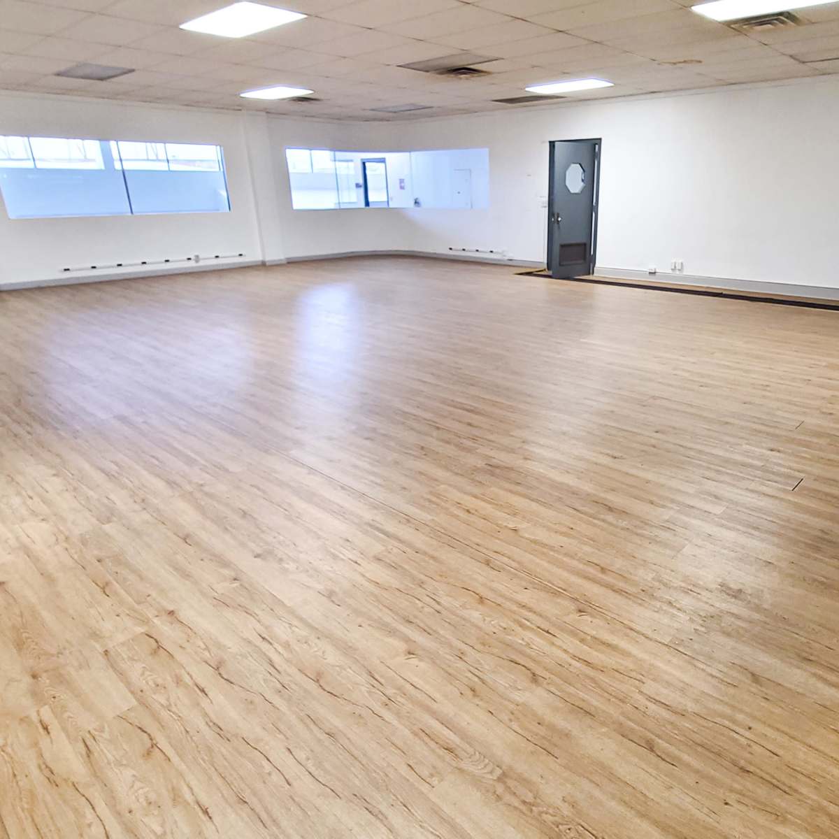 A spacious, empty room with wooden flooring and large windows that allow natural light to enter.
