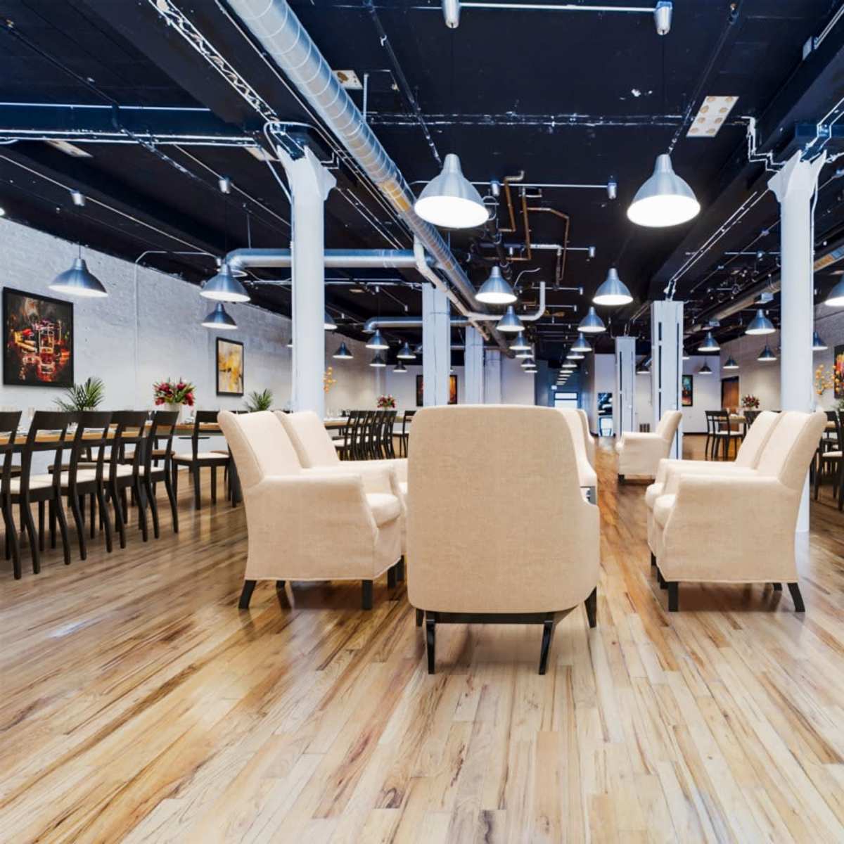 A spacious, well-lit event space with white walls, wooden floors, and rows of tables and chairs, along with armchairs arranged in the foreground.
