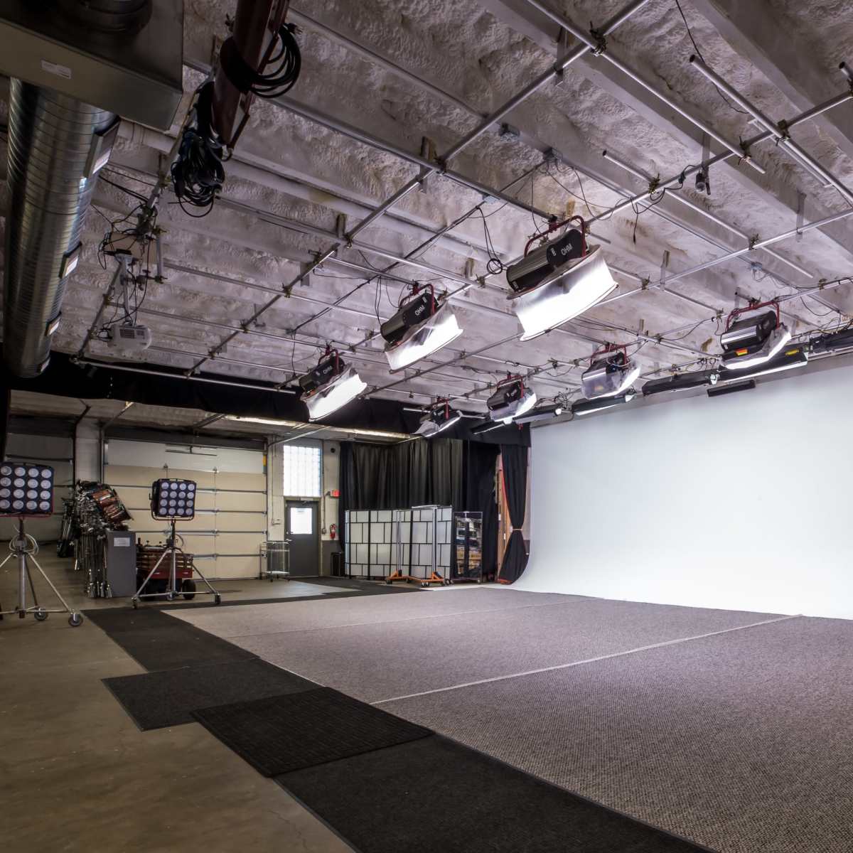 A photography studio with a large white backdrop, multiple lights mounted on the ceiling, and dark curtains along the sides.