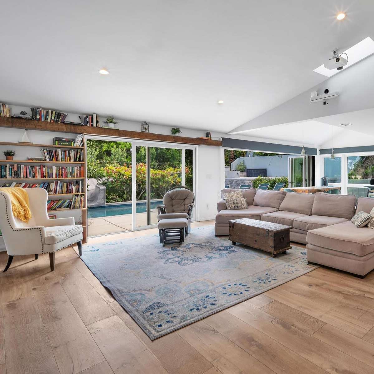 A modern living room features a large sectional sofa, a coffee table, bookshelves, and sliding glass doors that open to an outdoor patio.