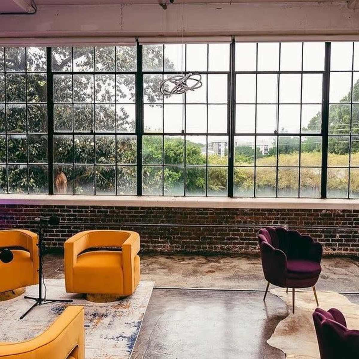The scene depicts a room with large, industrial-style windows, featuring yellow armchairs and a dark purple chair arranged on a rug next to a brick wall.