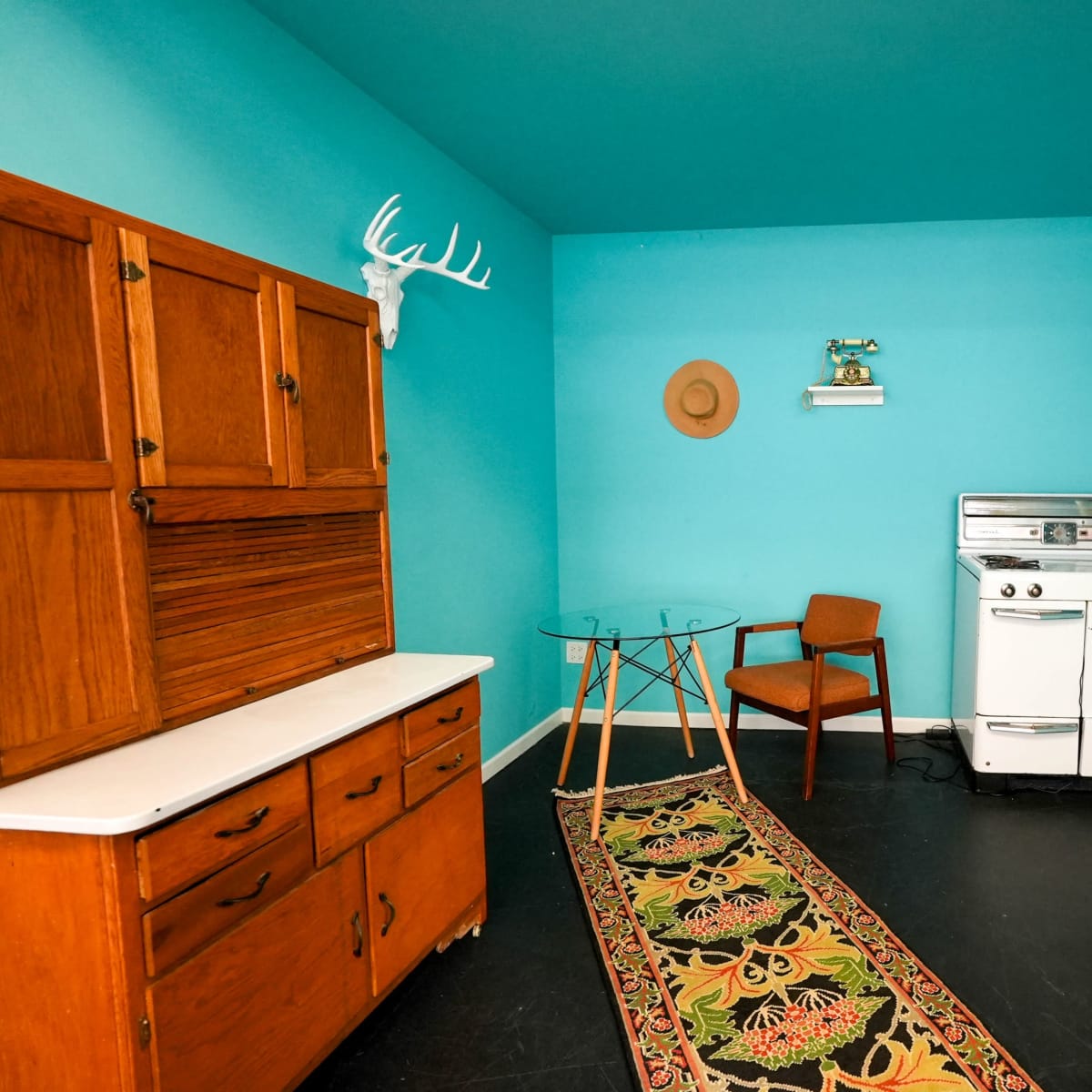 A teal-colored room with a wooden hutch, a glass dining table with a chair, an antique stove, and decorative wall-mounted antlers.