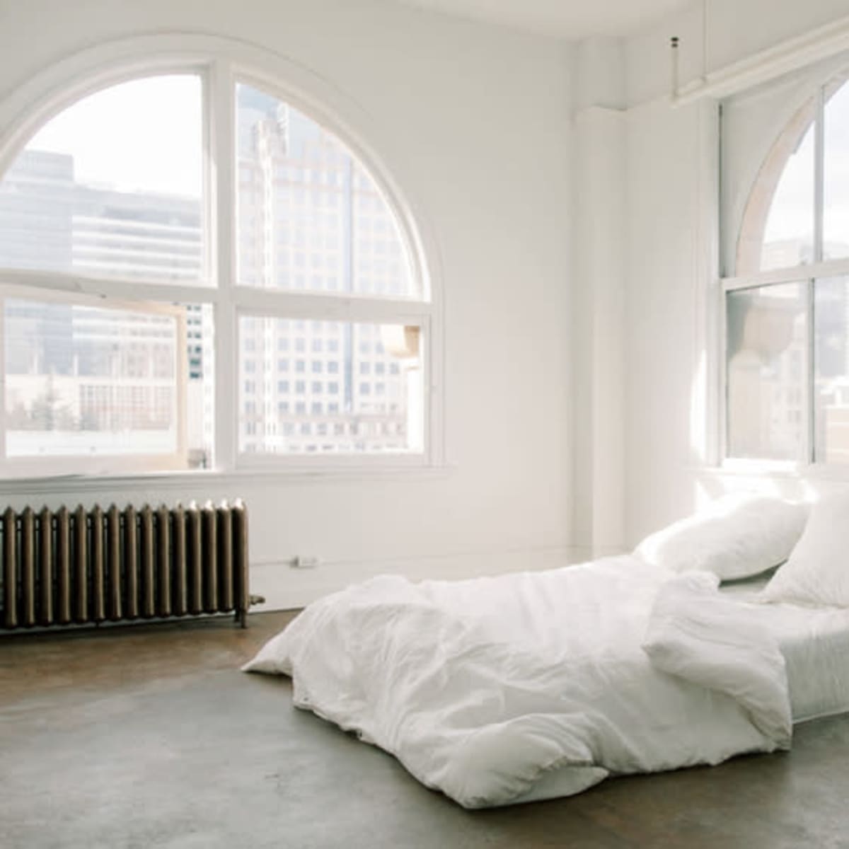 A minimalist bedroom features a unmade bed with white bedding in front of large arched windows overlooking a city skyline.