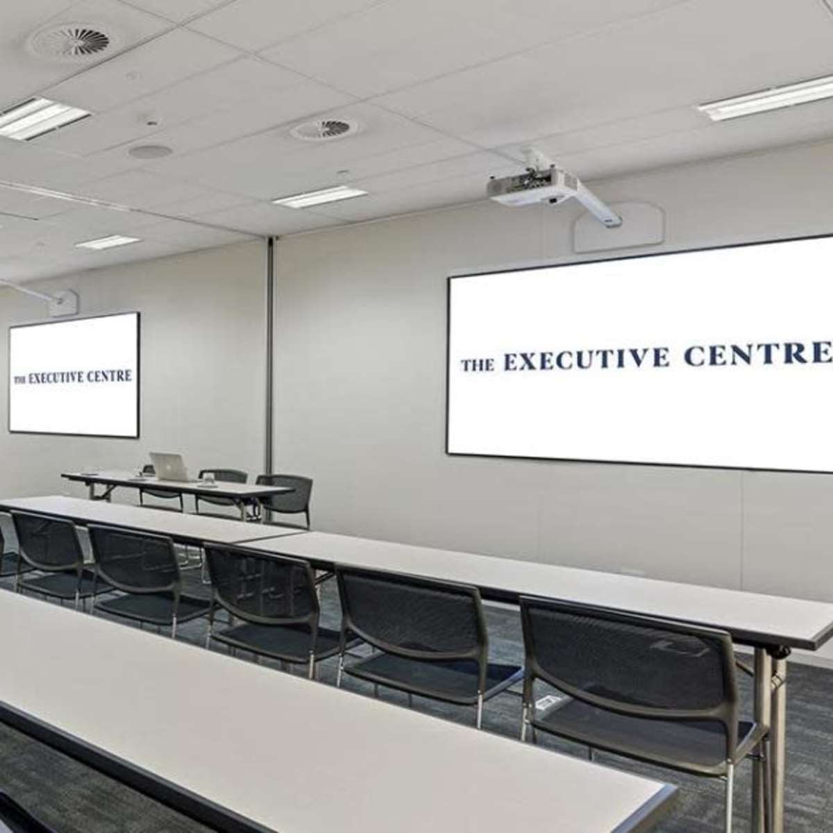 A modern conference room with long tables arranged in rows and two large screens displaying "THE EXECUTIVE CENTRE."