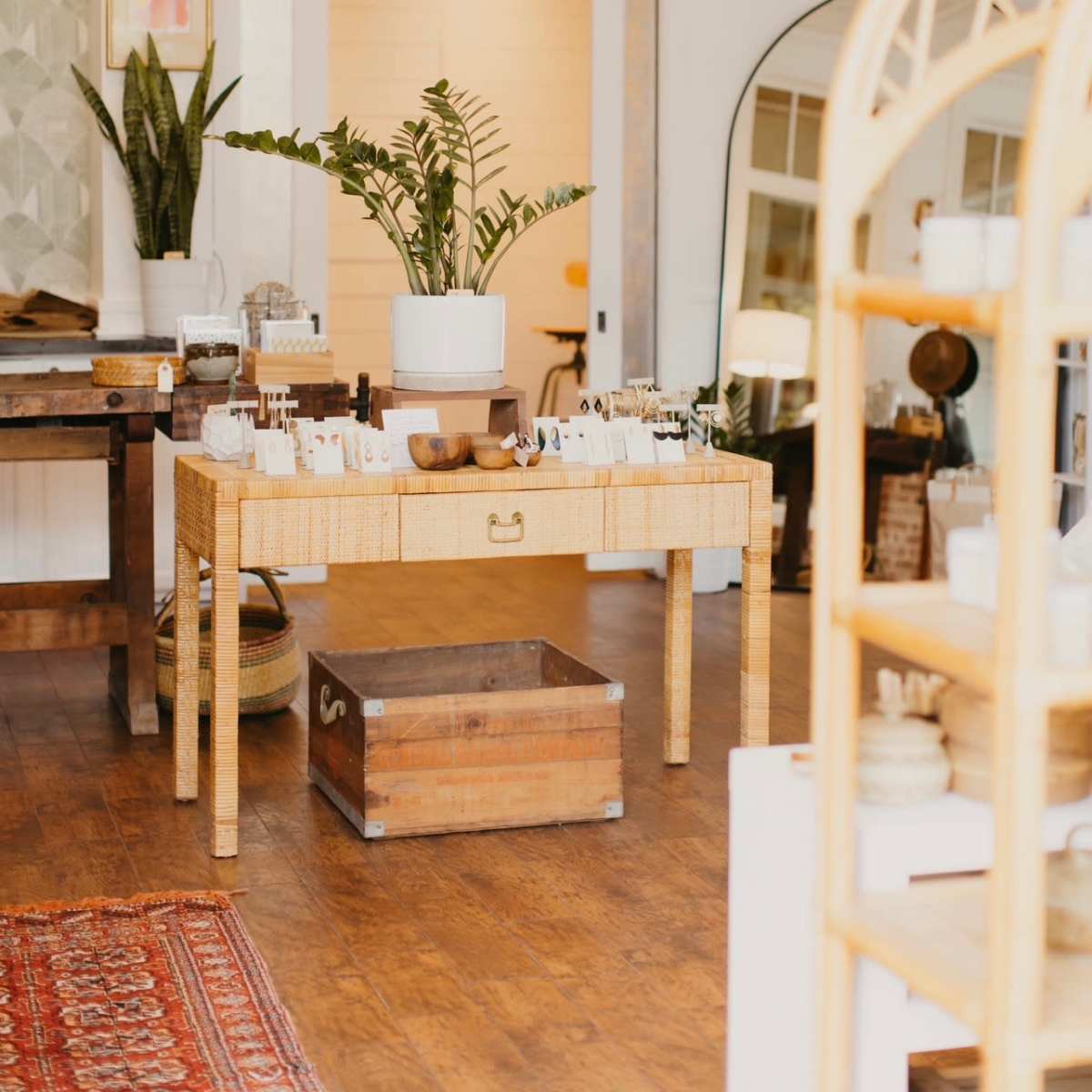 A well-organized retail space features a wooden table with various items for sale, surrounded by plants and decorative mirrors.