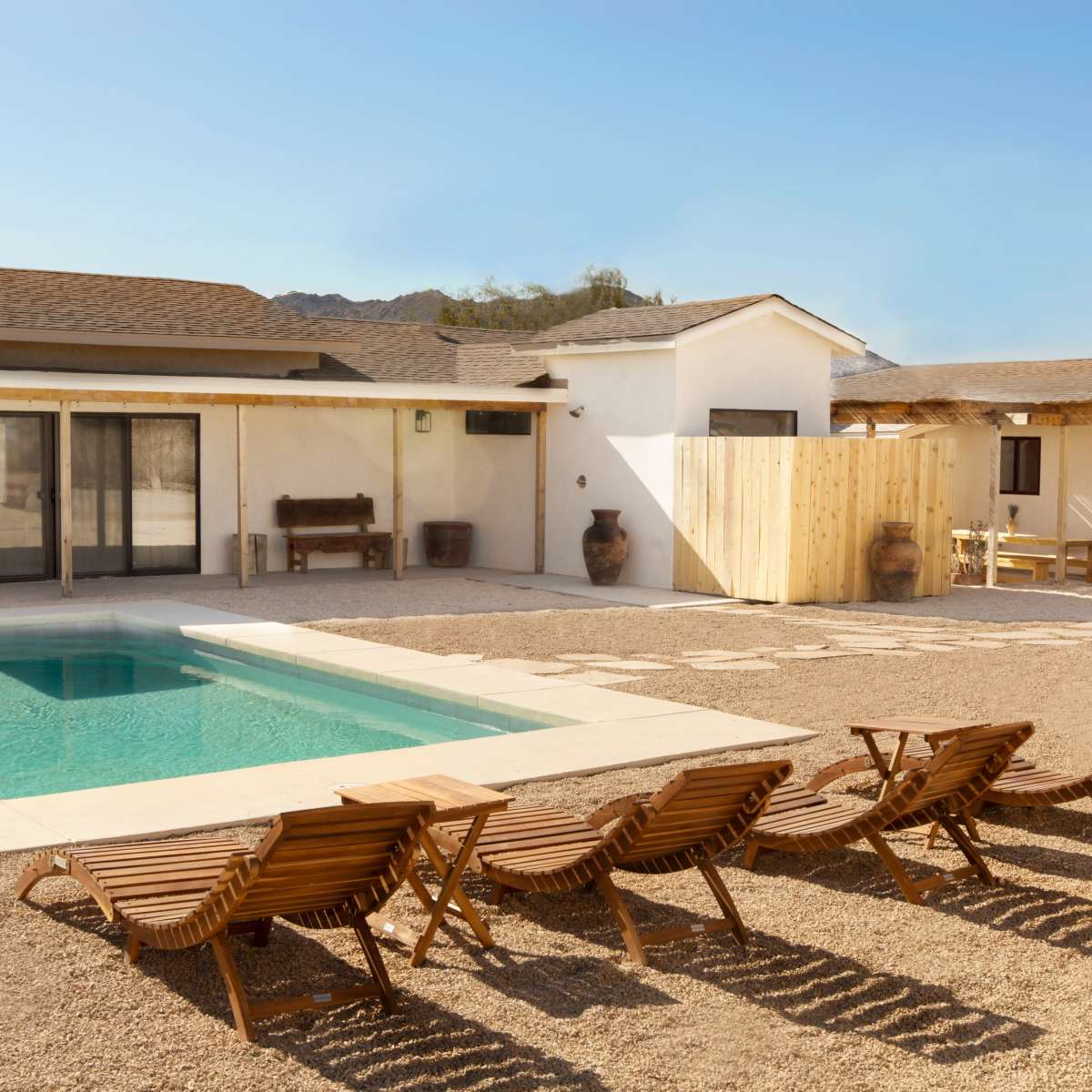 A modern white house with a pool and wooden lounge chairs is set in a gravel backyard surrounded by desert landscape.