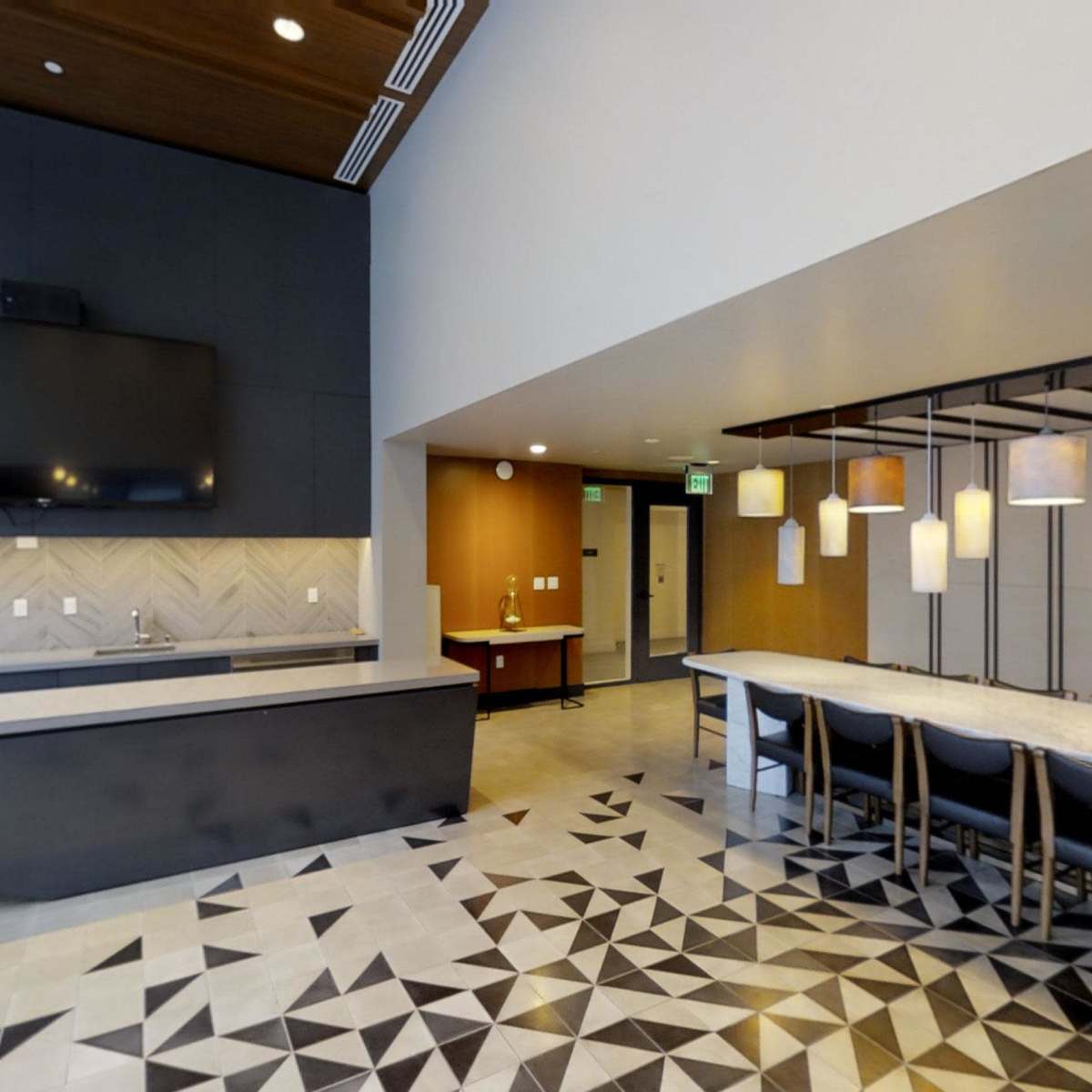 A modern communal kitchen area featuring a long table with chairs, a large television mounted on the wall, and pendant lighting above the table, complemented by a patterned floor.
