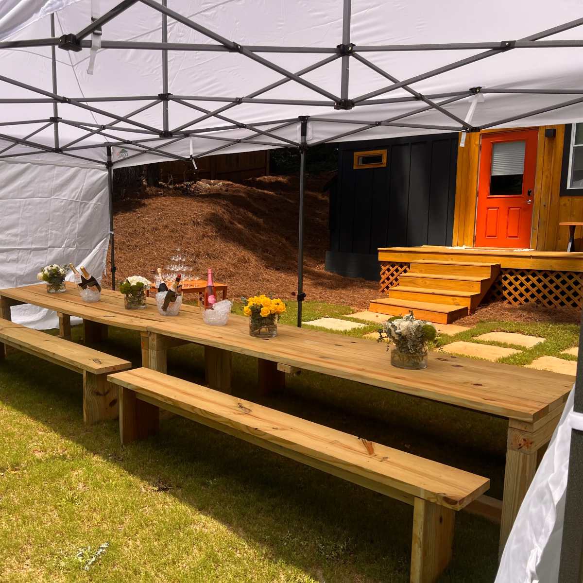 A wooden table with several vases of flowers is set up under a white canopy in a grassy area, leading to a house with an orange door.