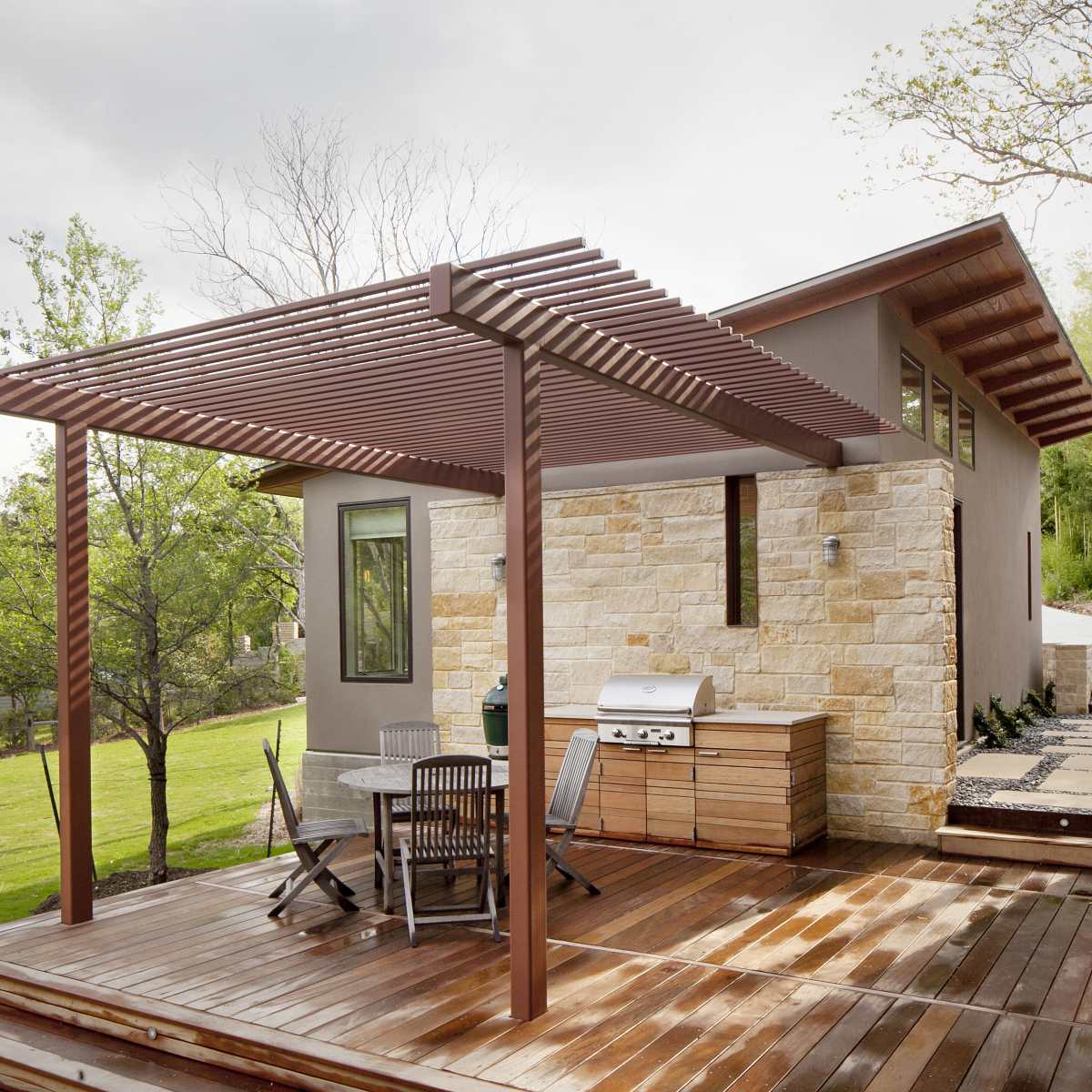 A modern wooden deck with a pergola, a grill area, and a stone-and-wood exterior of a house surrounded by greenery.
