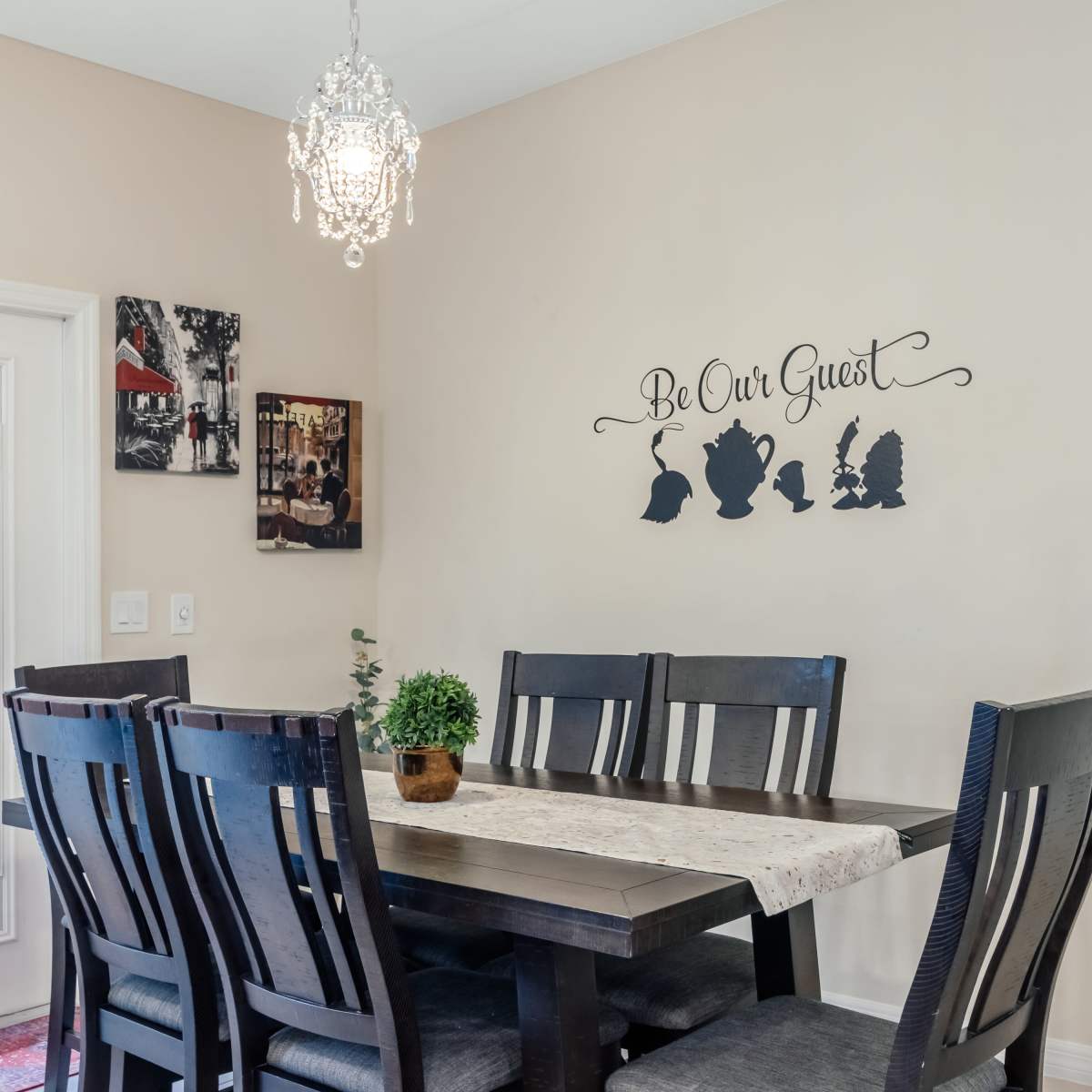 A dining area features a large table with eight chairs, a decorative wall sign, and a chandelier above.