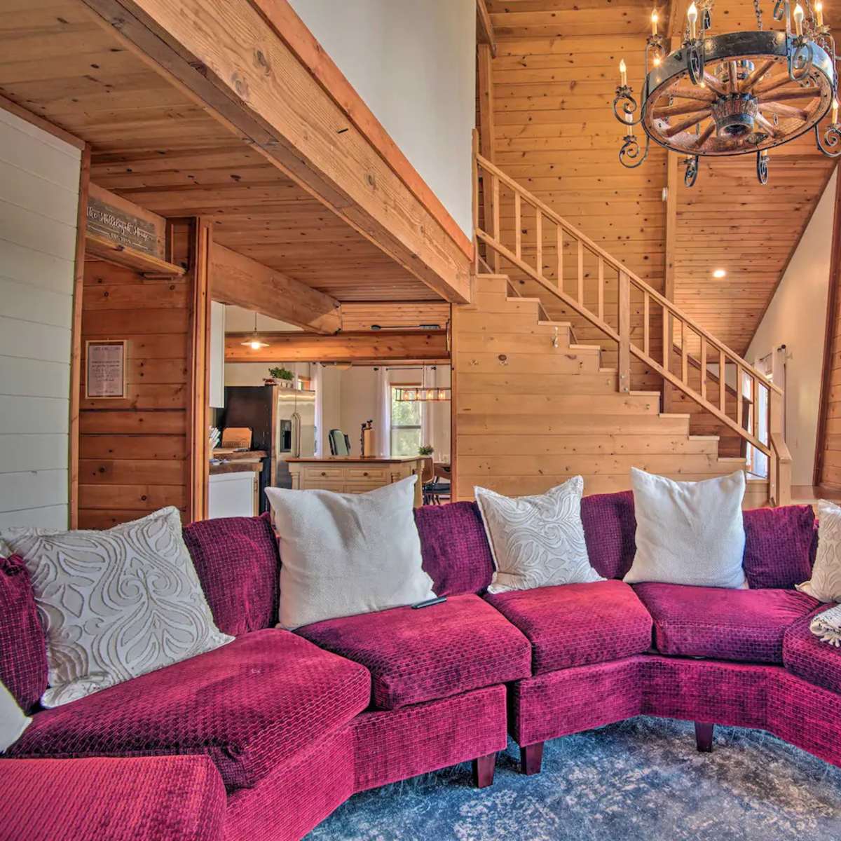 A cozy living room features a red sectional sofa with white pillows and large windows, allowing natural light to illuminate the wooden interior and staircase.