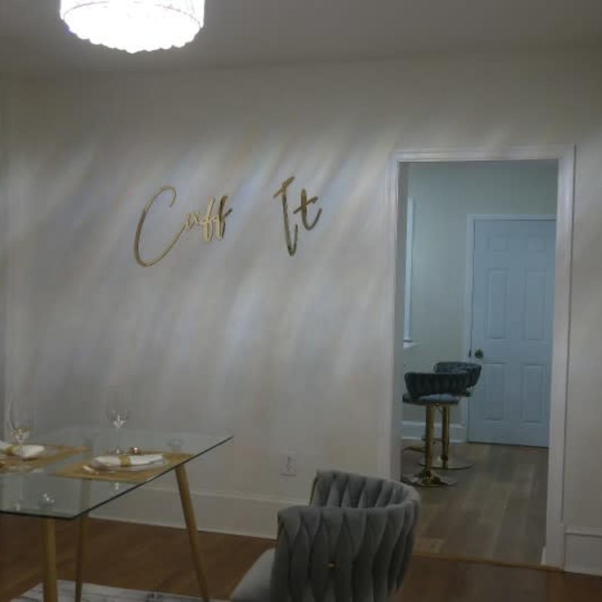 A modern dining area features a glass table set for two, surrounded by tufted gray chairs, with decorative gold wall art and a mirror enhancing the space.