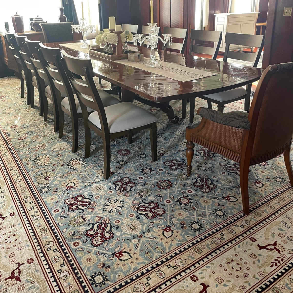 A spacious dining room with a long wooden table set for dinner, surrounded by chairs and featuring ornate rugs and curtains.