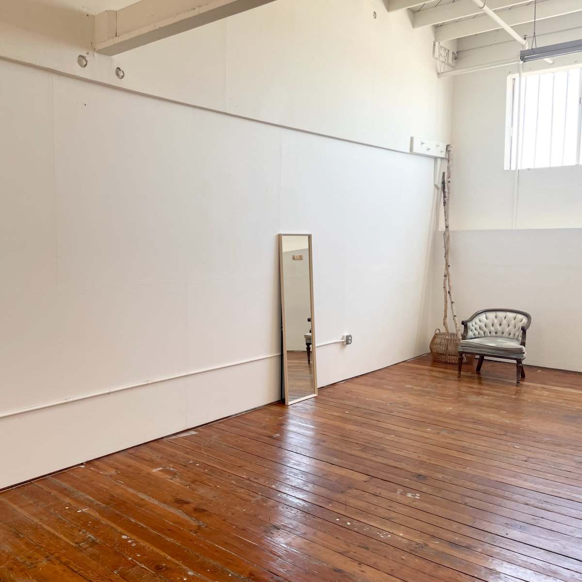 A spacious room with white walls, a wooden floor, a tall mirror leaning against the wall, and a vintage chair.