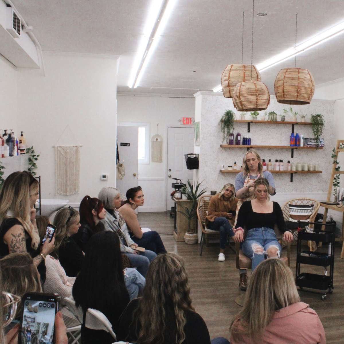 A group of people sits in a salon, watching a stylist demonstrate hair techniques on a model in a chair.