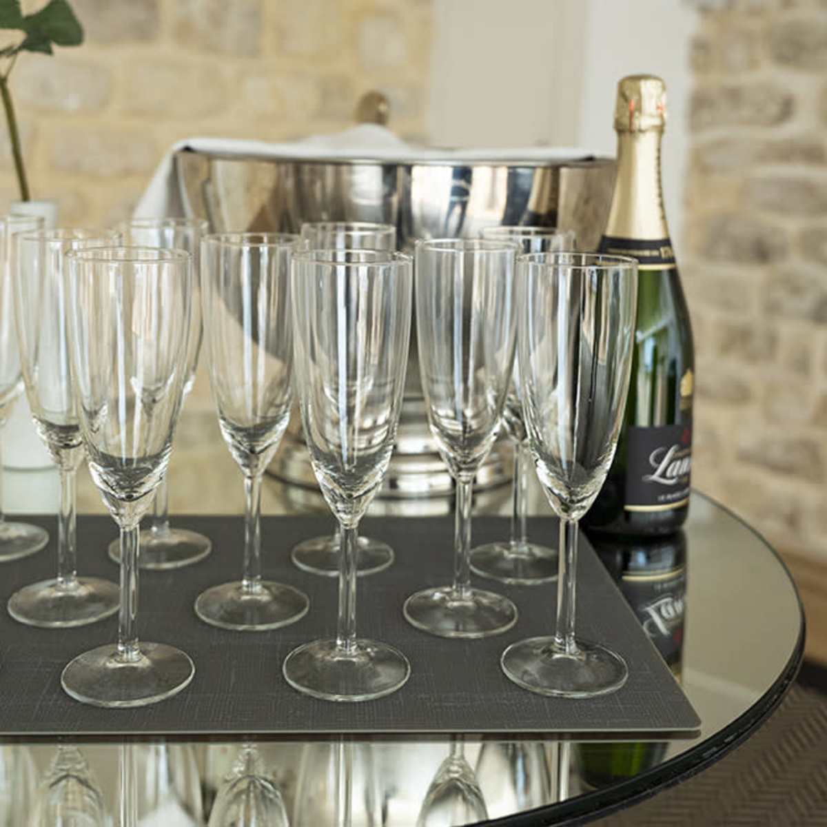 A set of champagne flutes is arranged on a mirrored table next to a bottle of champagne and an ice bucket, against a stone wall backdrop.