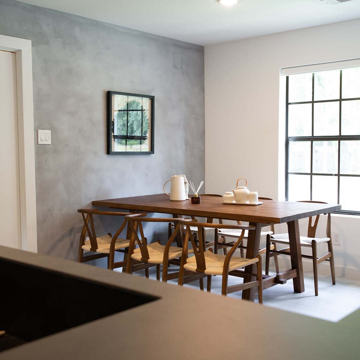 A modern dining area features a wooden table surrounded by chairs, with a minimalistic decor against a gray wall and large windows providing natural light.
