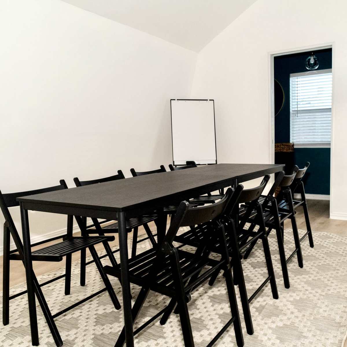 A long black table surrounded by eight folding chairs is set in a light-colored room with an open door leading to a blue-walled space.