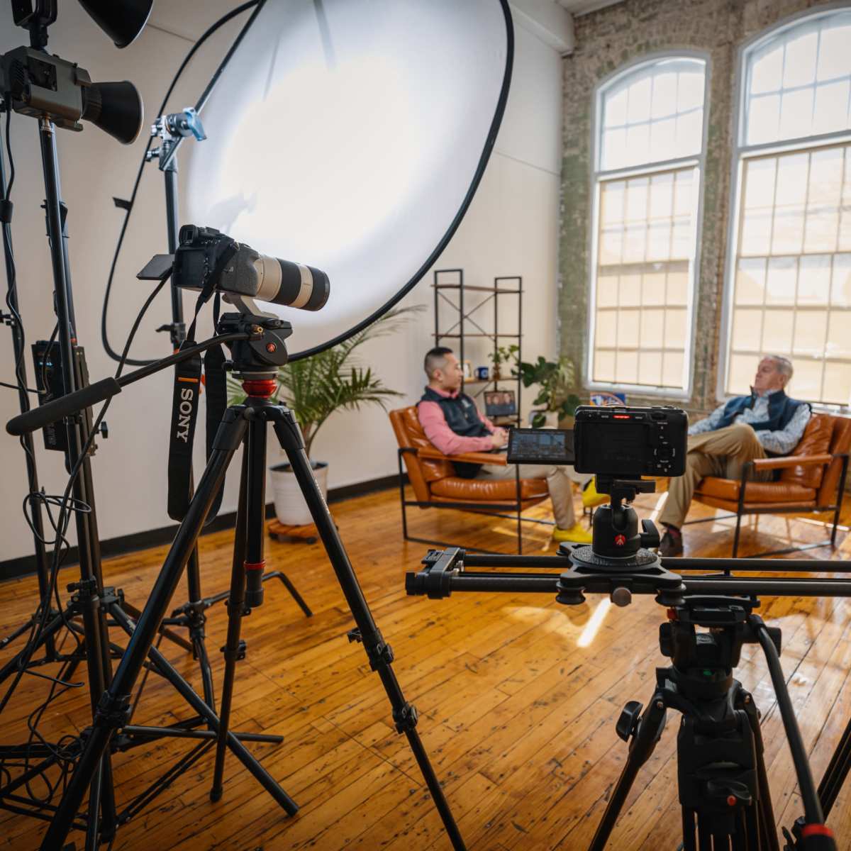 A film crew sets up equipment in a spacious, well-lit room to record an interview between two men sitting on leather chairs.