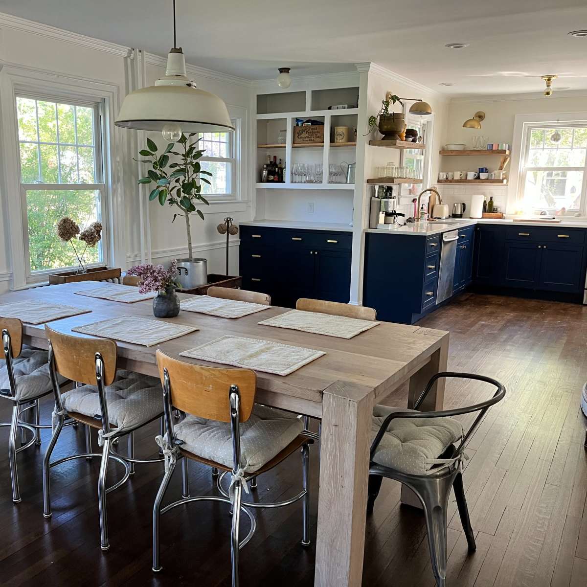 A modern dining area with a large wooden table surrounded by six chairs, set adjacent to a kitchen featuring blue cabinets and ample natural light.