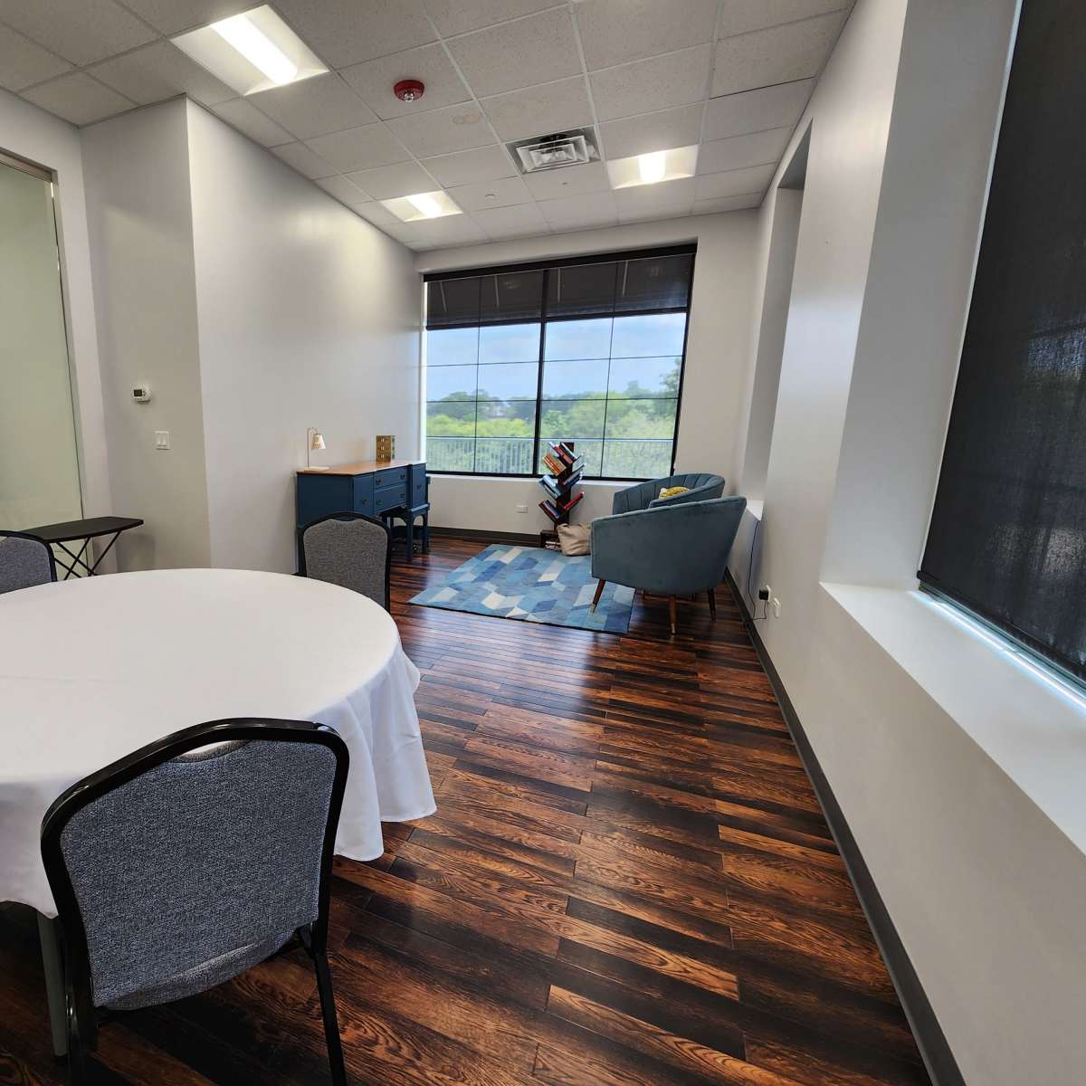 A bright, modern room with a round table covered by a white tablecloth, a blue armchair, and a large window overlooking greenery.