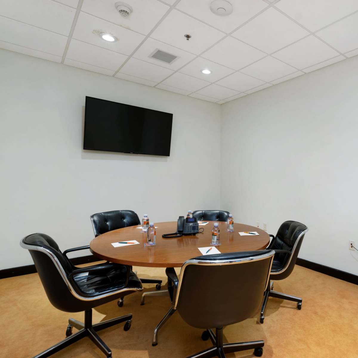 A small conference room features a round wooden table surrounded by four black leather chairs, a wall-mounted TV, a printer, and water bottles on the table.