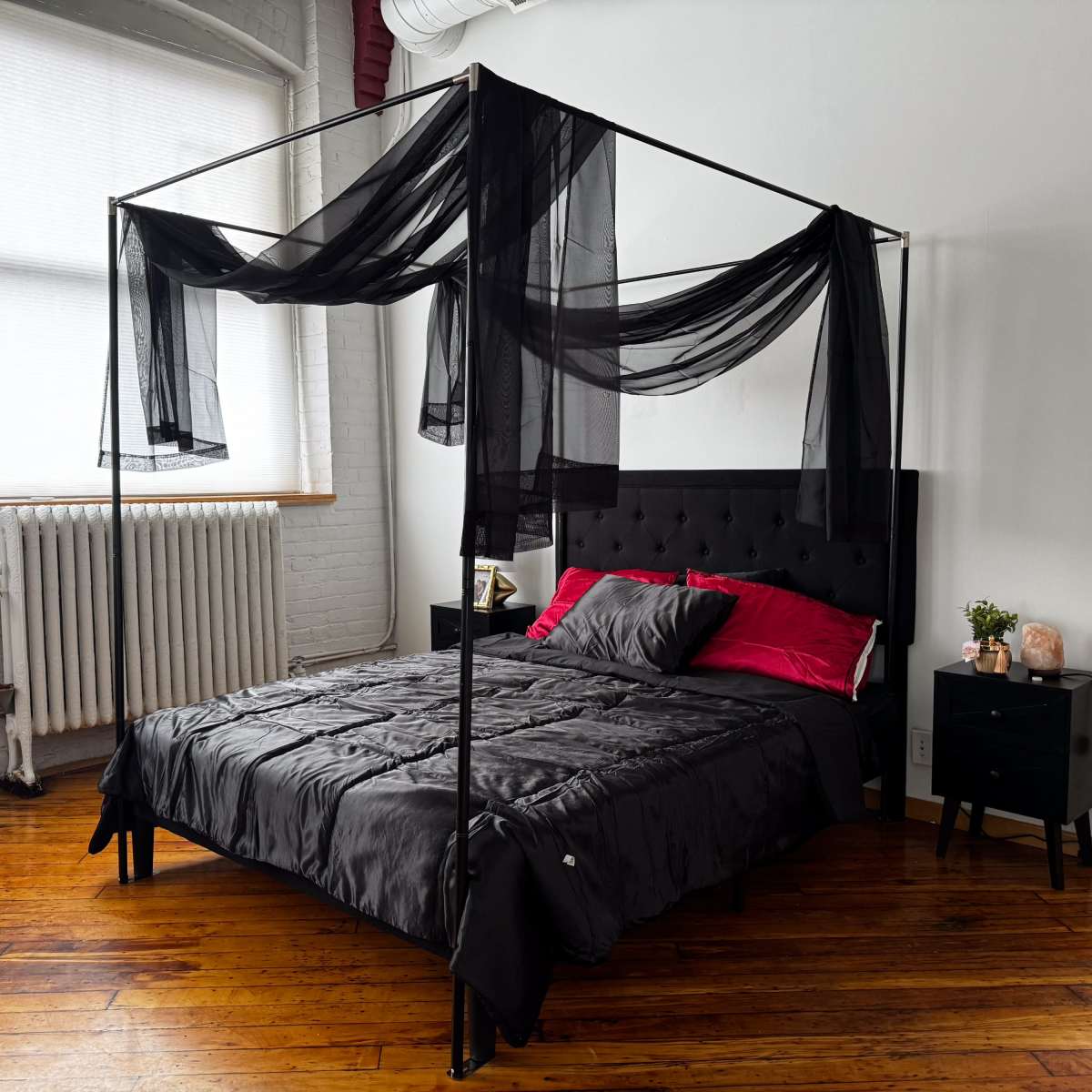 A canopy bed with black drapery, red pillows, a dark quilt, and a nightstand beside it in a room with wooden flooring and a bright window.