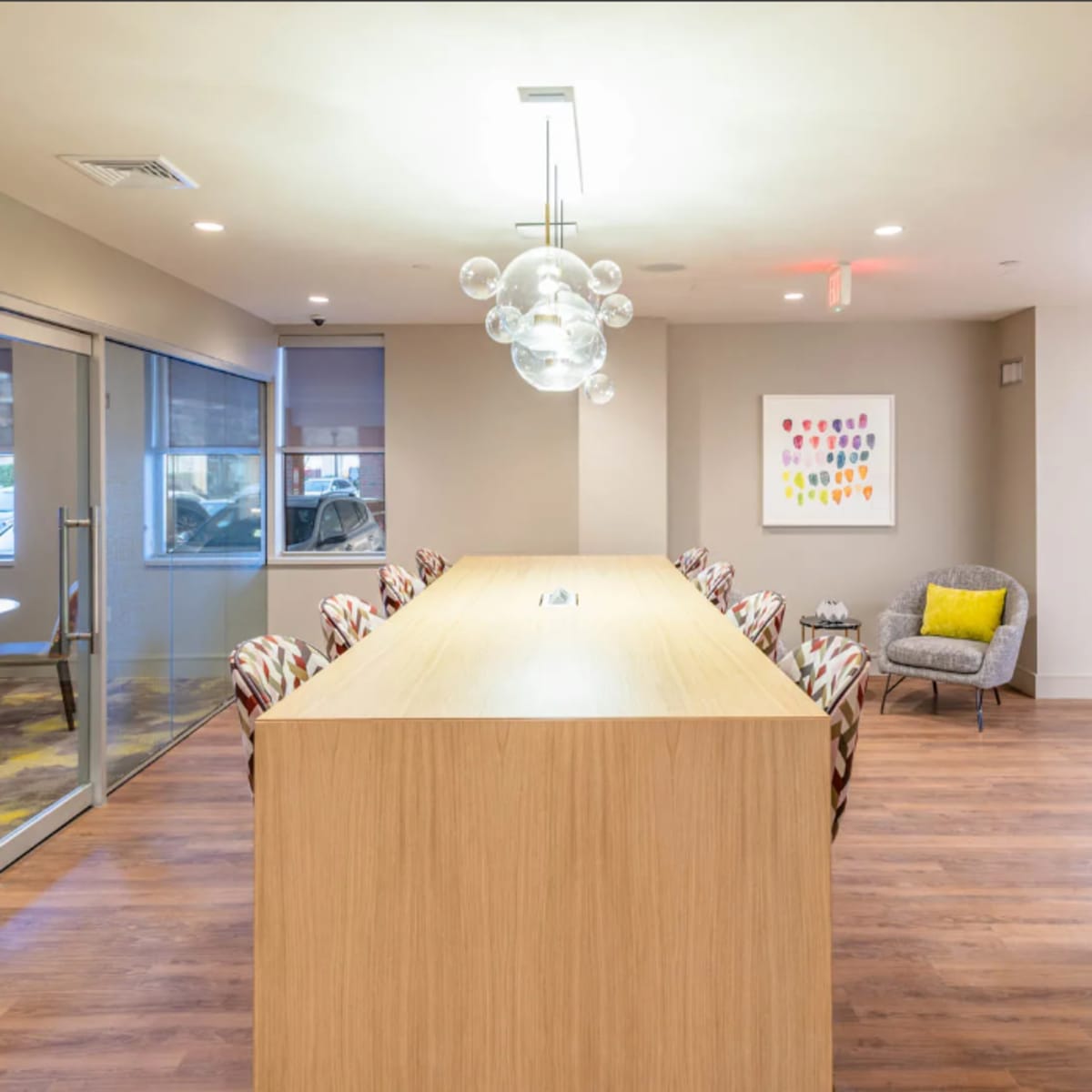 A modern office space features a long wooden table with seating and a seating area with a gray chair and colorful artwork on the wall.