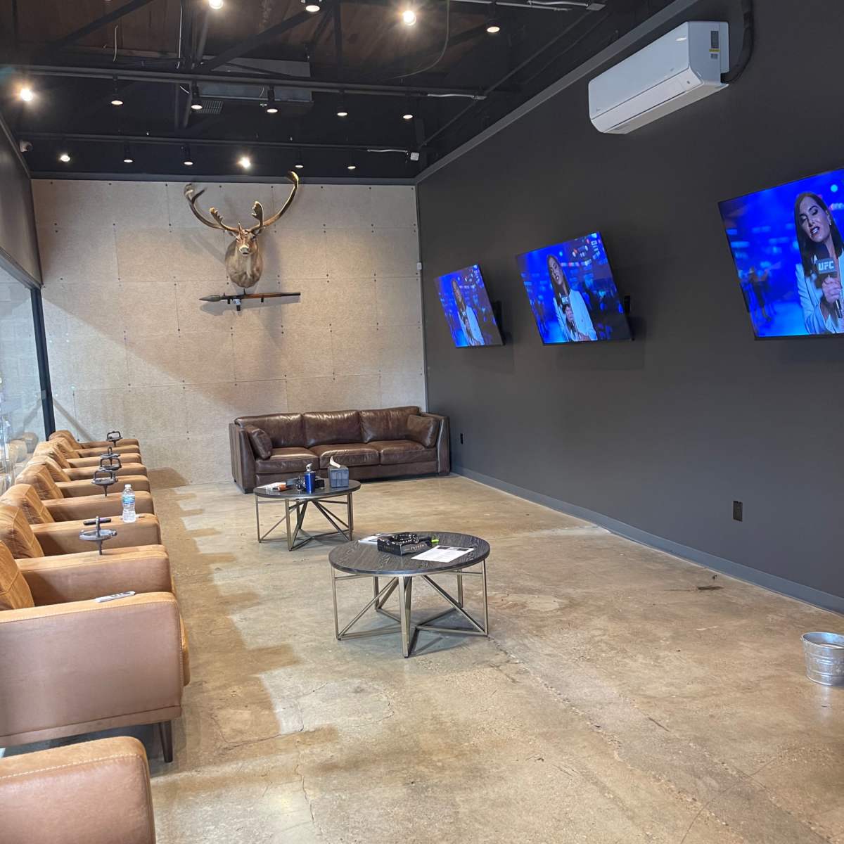 A modern lounge area with brown leather seating, three flat-screen TVs on the black wall, and a mounted deer head above a concrete section.