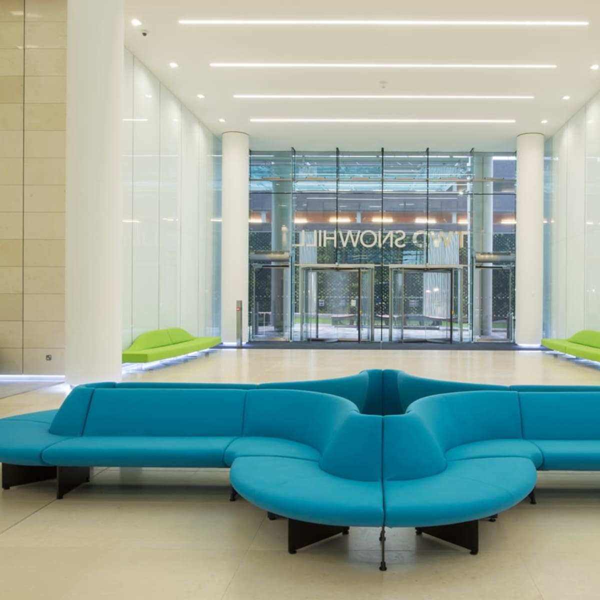 A spacious lobby featuring a large, stylized blue sofa in the center and two green sofas along the walls, with glass doors in the background.