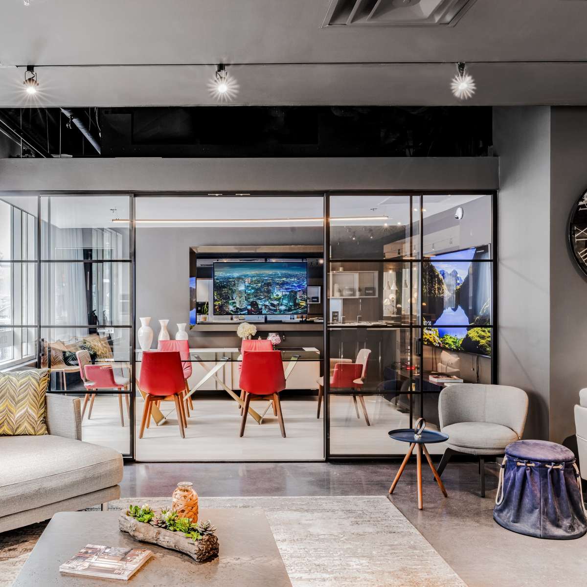 A modern living area features a gray sectional sofa, a glass partition leading to a dining space with red chairs, and contemporary decor elements throughout.