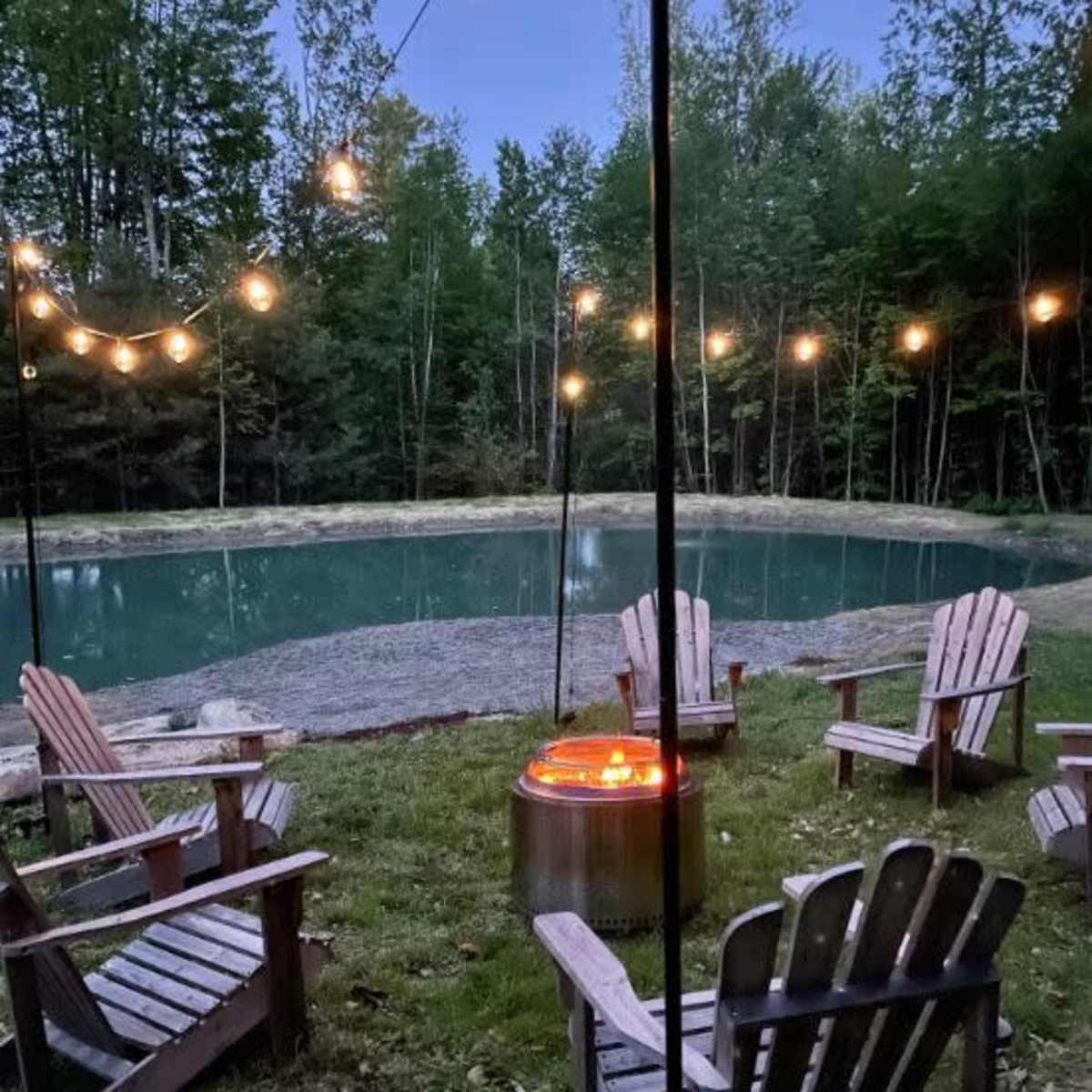 A circle of Adirondack chairs surrounds a fire pit near a pond, illuminated by string lights in a wooded area.