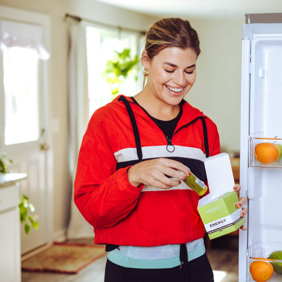 A woman in a bright red jacket is smiling while holding a box labeled "ENERGY" in front of an open refrigerator filled with juice boxes and apples.