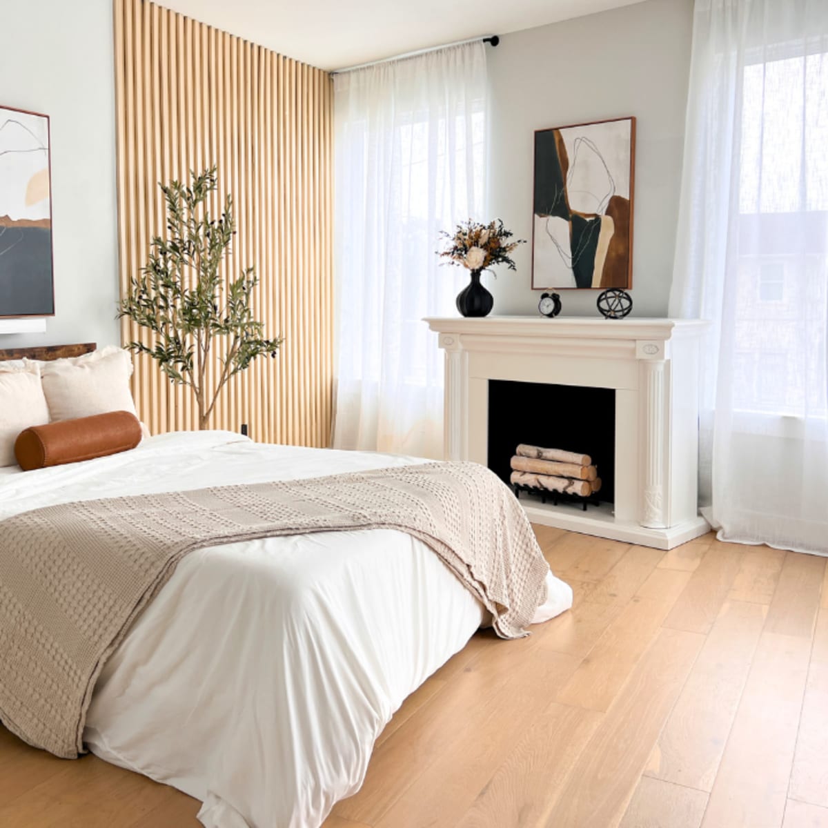 A well-decorated bedroom features a bed with white linens, a textured throw, a wooden headboard, and a fireplace surrounded by minimalist artwork and natural decor.